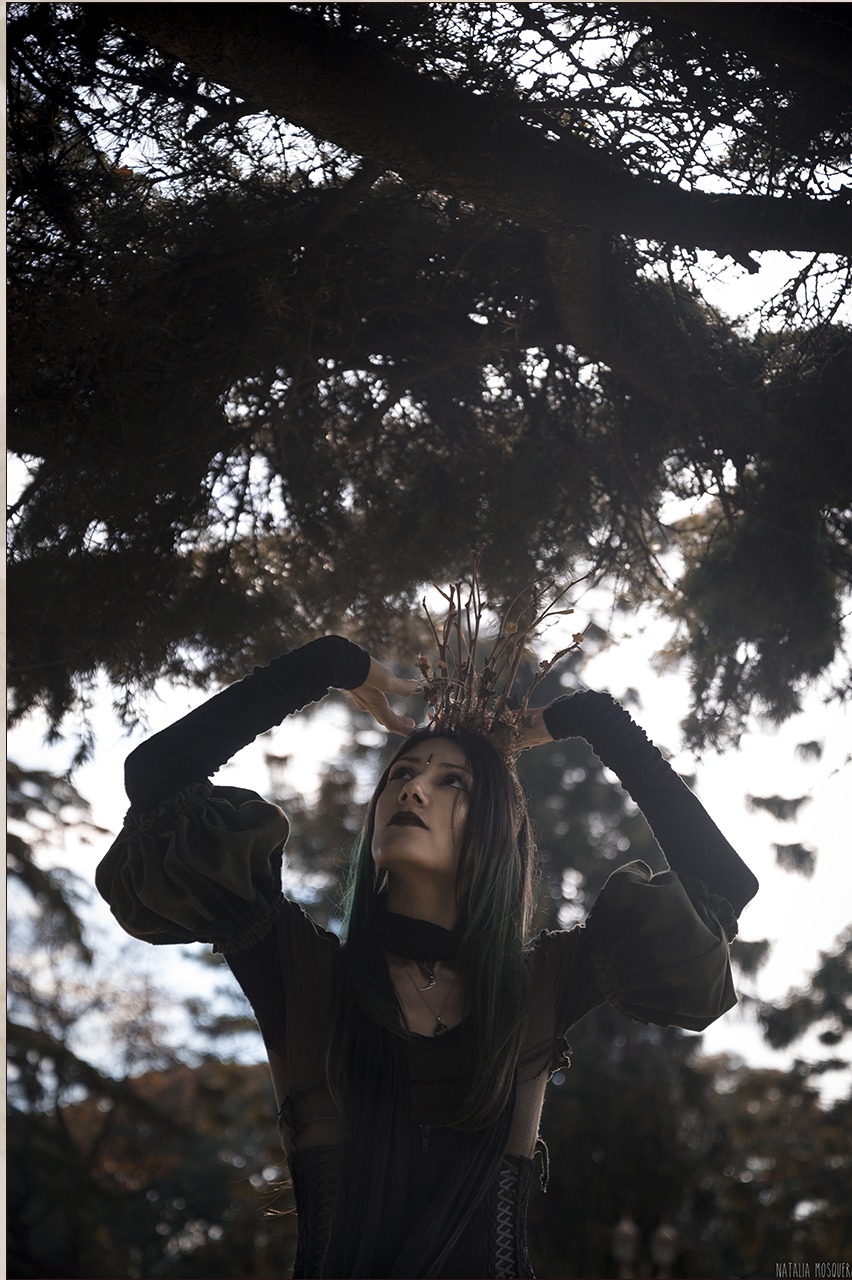 Carla standing in a forest, looking up through branches, wearing a twig crown